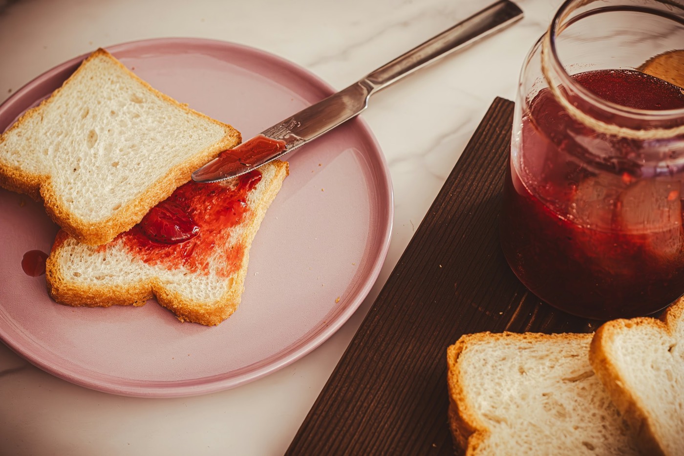 Toast spread with strawberry jam on a pink plate, beside an open jar of jam