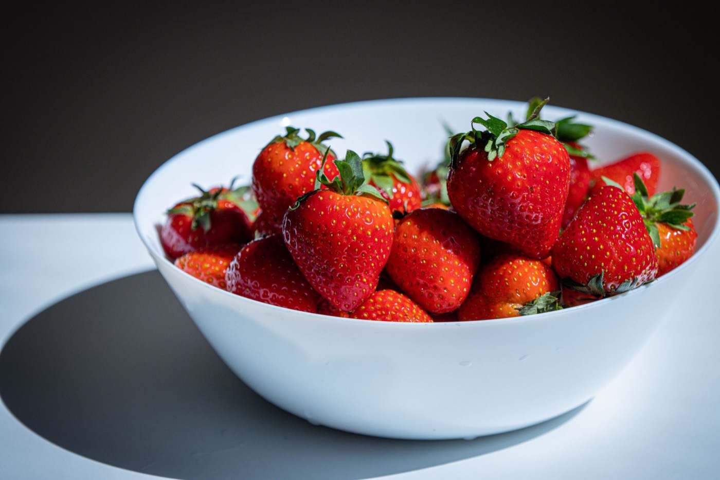 A white bowl heaped with whole ripe strawberries in bright sunlight